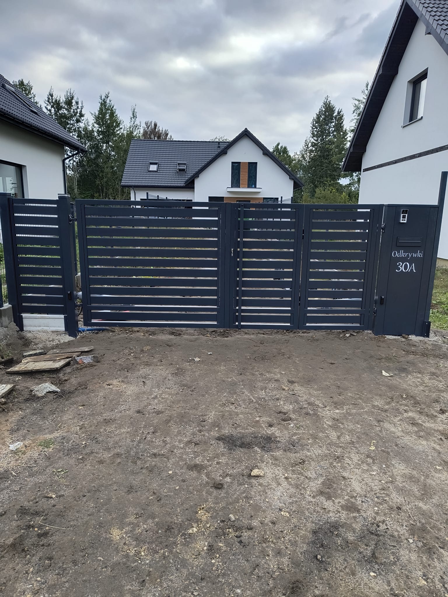Nowoczesna, antracytowa brama wjazdowa z poziomymi panelami, z adresem 'Odkrywki 30A', na tle domów jednorodzinnych i pochmurnego nieba.