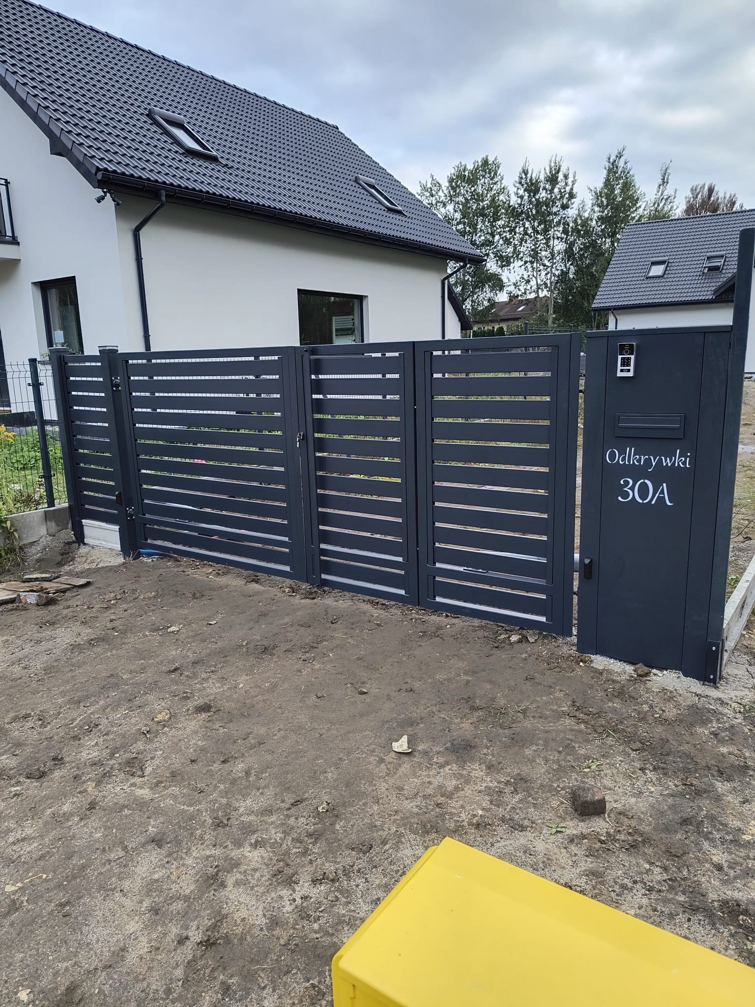 Nowoczesna, pozioma brama wjazdowa w kolorze grafitowym z numerem domu 'Odkrywki 30A', dom w tle. Minimalistyczny design z domofonem.