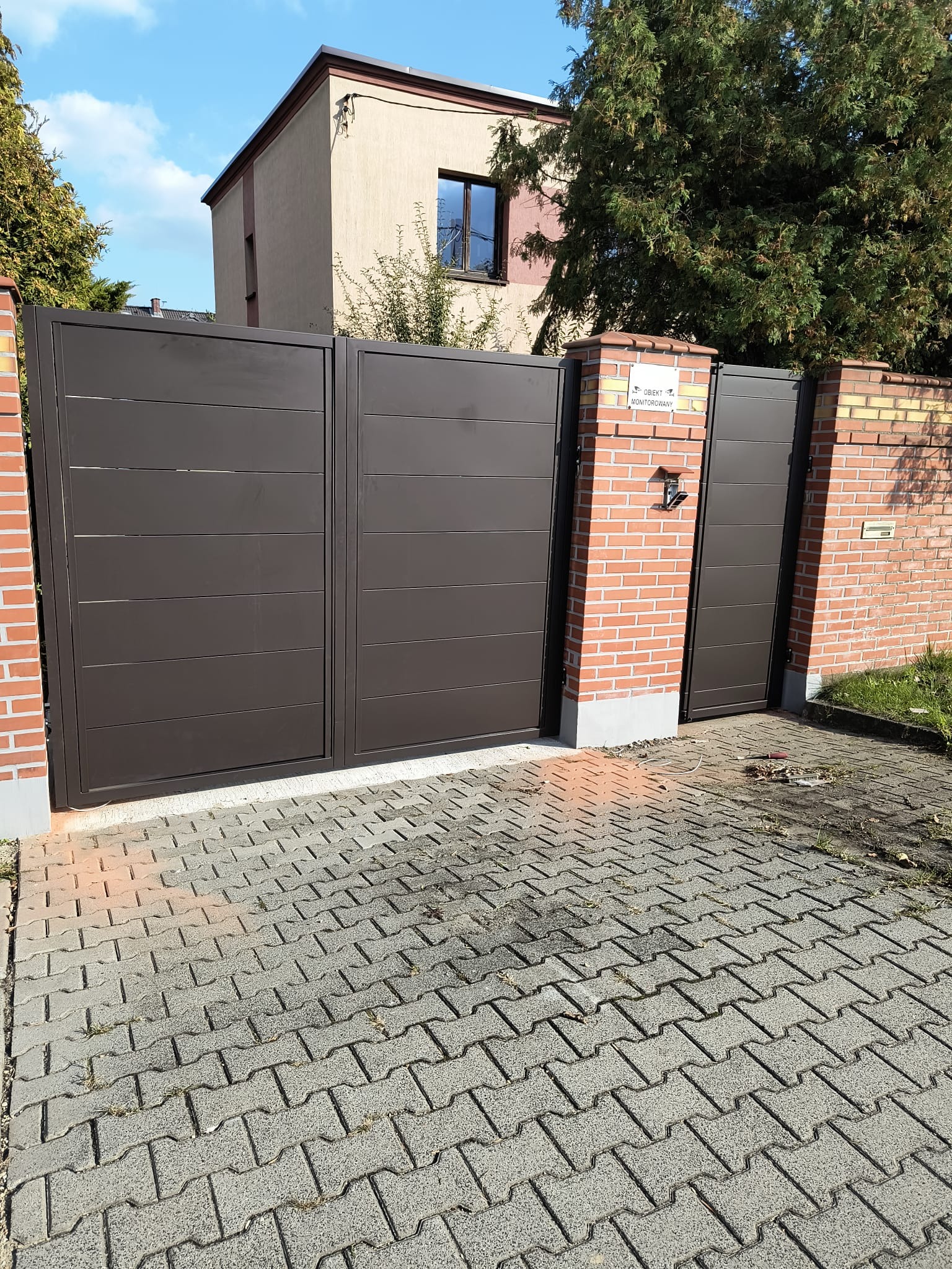 Nowoczesna, brązowa brama wjazdowa z poziomymi panelami, zamontowana na murze z cegły. Widoczny dom w tle i kostka brukowa na podjeździe.
