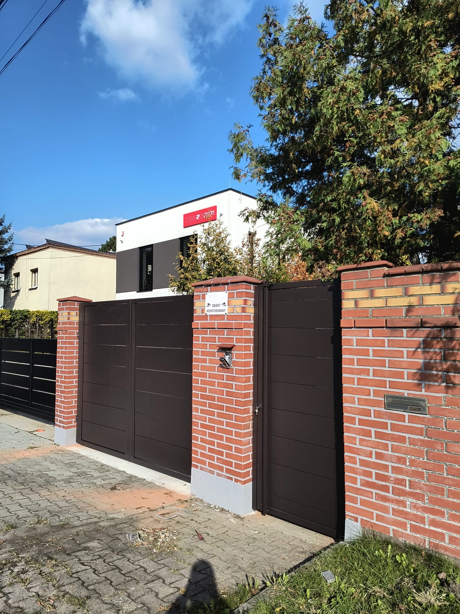 Nowoczesna brama wjazdowa w kolorze brązowym z ceglanymi słupkami i furtką. Widoczny znak 'obiekt monitorowany' i skrzynka na listy. Błękitne niebo w tle.