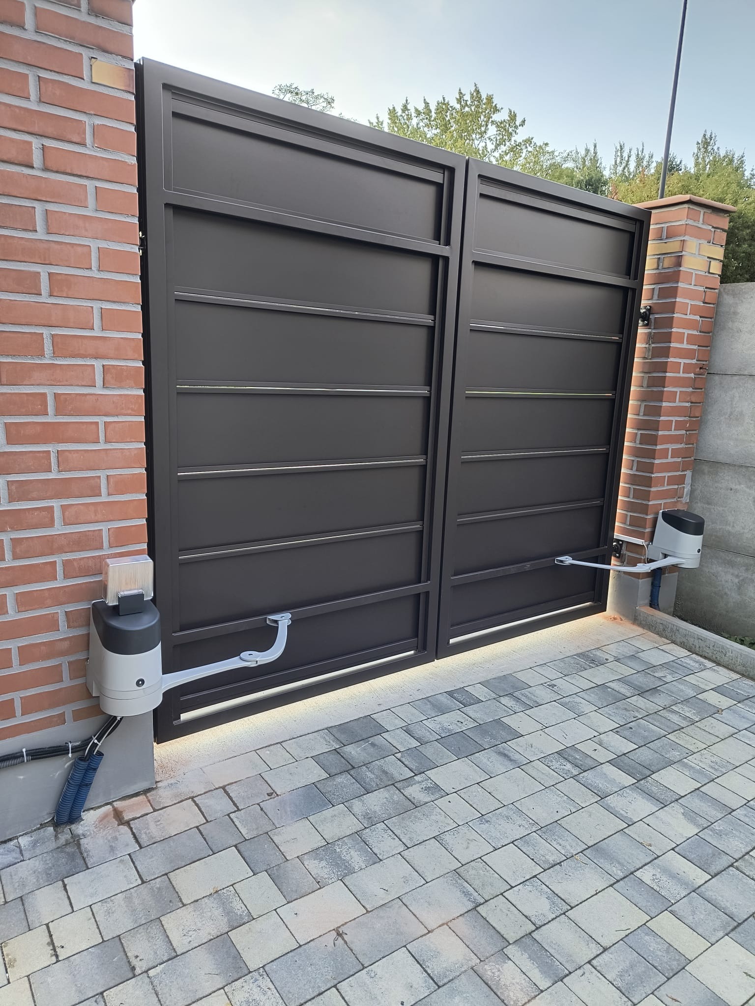 Nowoczesna, dwuskrzydłowa brama wjazdowa w kolorze antracytowym z automatyką, zamontowana na ceglanych słupkach. Widoczna kostka brukowa i zieleń w tle.