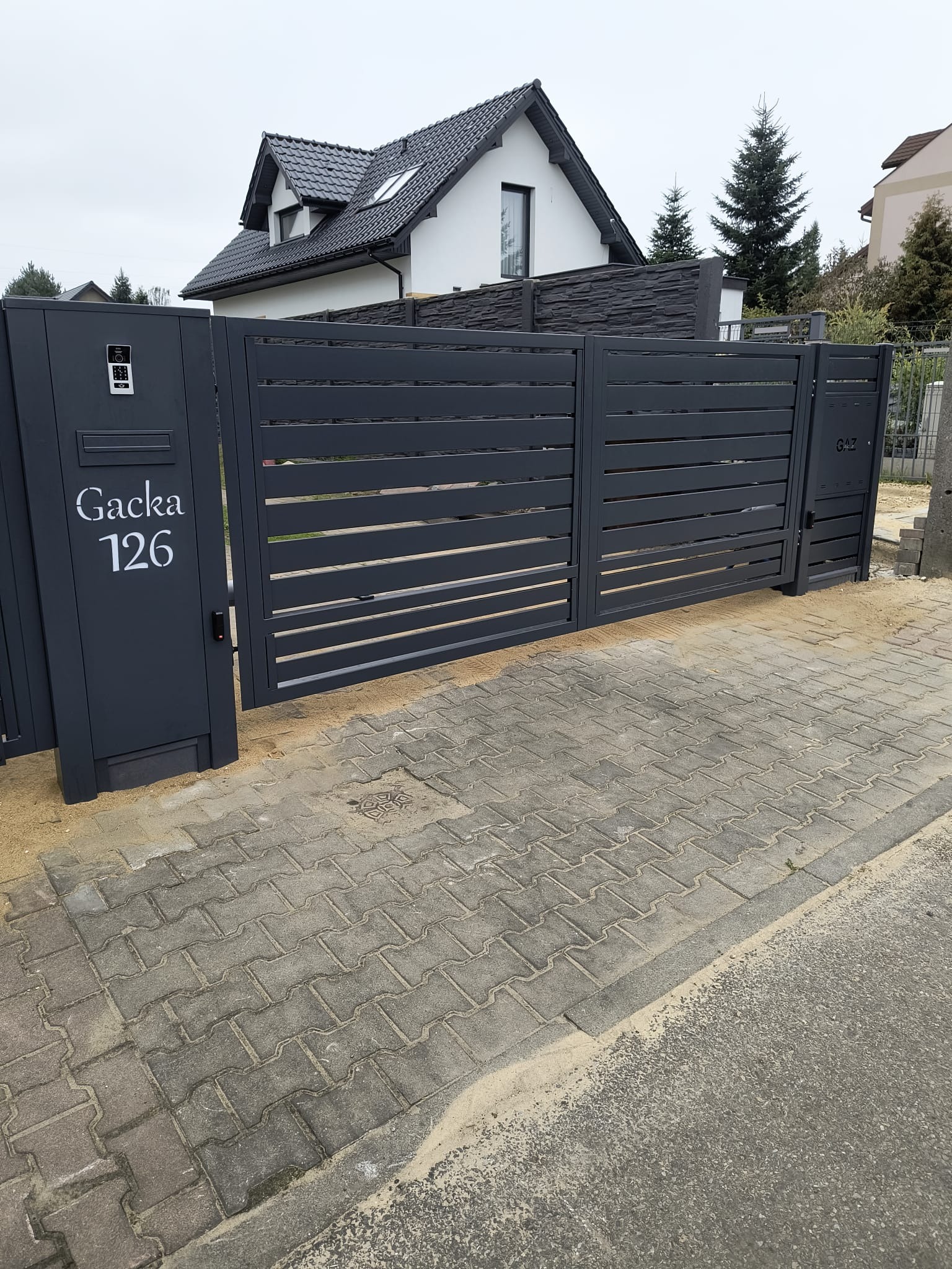 Nowoczesna brama wjazdowa z szarego metalu, z numerem domu 'Gacka 126' i domofonem, na tle kostki brukowej i domu z ciemnym dachem.