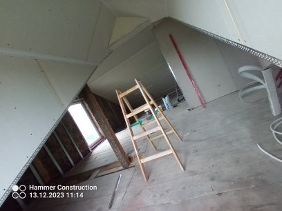 Montaż płyt kartonowo-gipsowych na poddaszu, widoczna drewniana drabina, surowe drewno i okno dachowe, etap prac wykończeniowych.