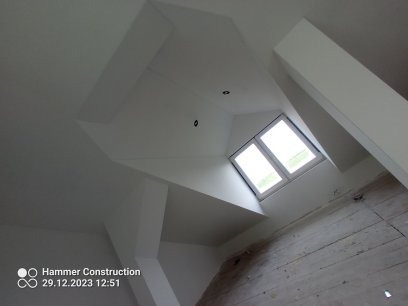 Białe wnętrze poddasza z oknem dachowym, widoczne płyty karton gips na skosach i fragment drewnianej podłogi w trakcie remontu.