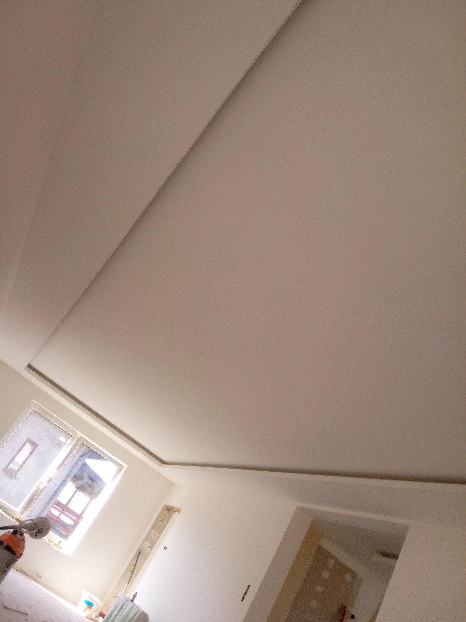 Wykończony sufit podwieszany z płyt kartonowo-gipsowych w jasnym pomieszczeniu, widok ukośny z elementami okna i drzwi w tle.