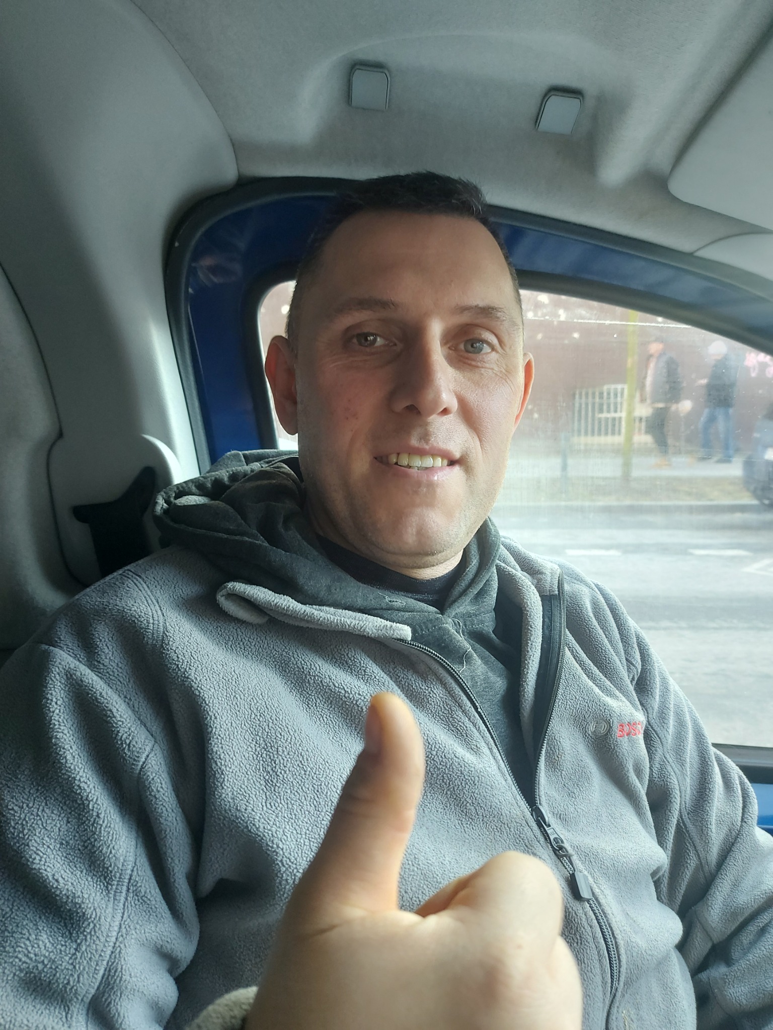 Uśmiechnięty mężczyzna w szarej bluzie z logo BOSCH, siedzący w samochodzie, pokazuje kciuk w górę; widok przez okno na ulicę.