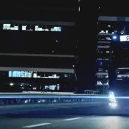 Ciężar&oacute;wka z naczepą marki Volvo z napisem SB ROAD na tle nocnego krajobrazu miejskiego z biurowcami i rozmazanymi światłami przejeżdżających samochod&oacute;w.