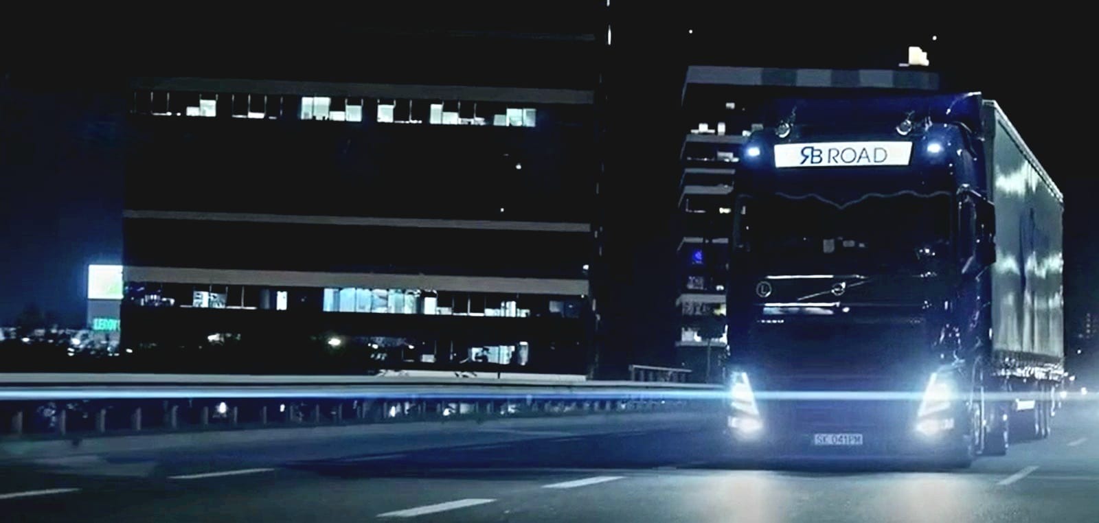 Ciężarówka z naczepą marki Volvo z napisem SB ROAD na tle nocnego krajobrazu miejskiego z biurowcami i rozmazanymi światłami przejeżdżających samochodów.