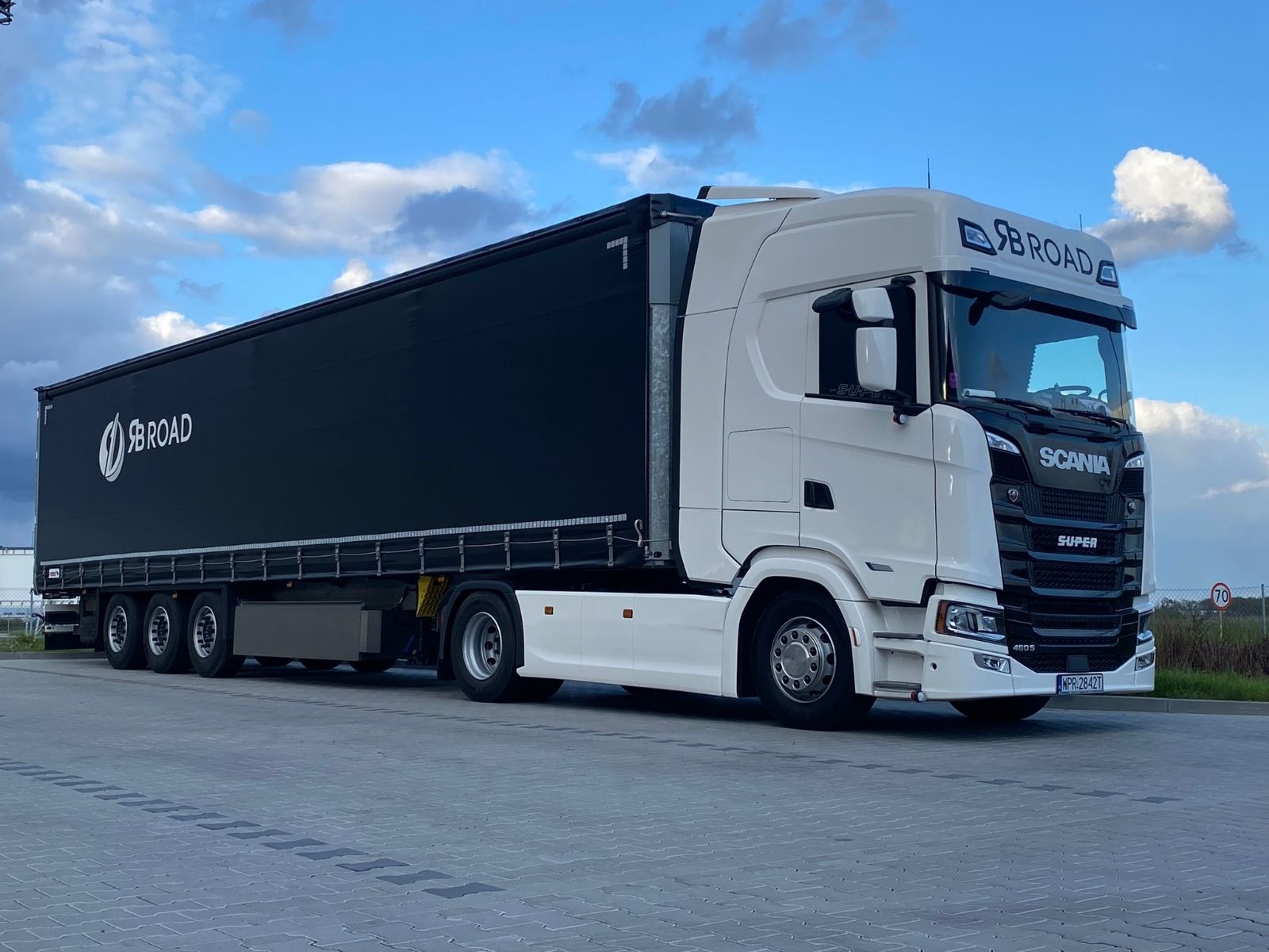 Biała ciężarówka Scania z czarną naczepą z logo firmy SB ROAD, zaparkowana na betonowym placu pod błękitnym niebem z chmurami.