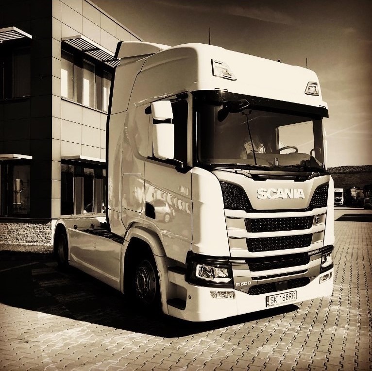 Ciągnik siodłowy Scania R500 w kolorach sepii, zaparkowany przed nowoczesnym budynkiem biurowym z widoczną tablicą rejestracyjną SK 168RR, na kostce brukowej.