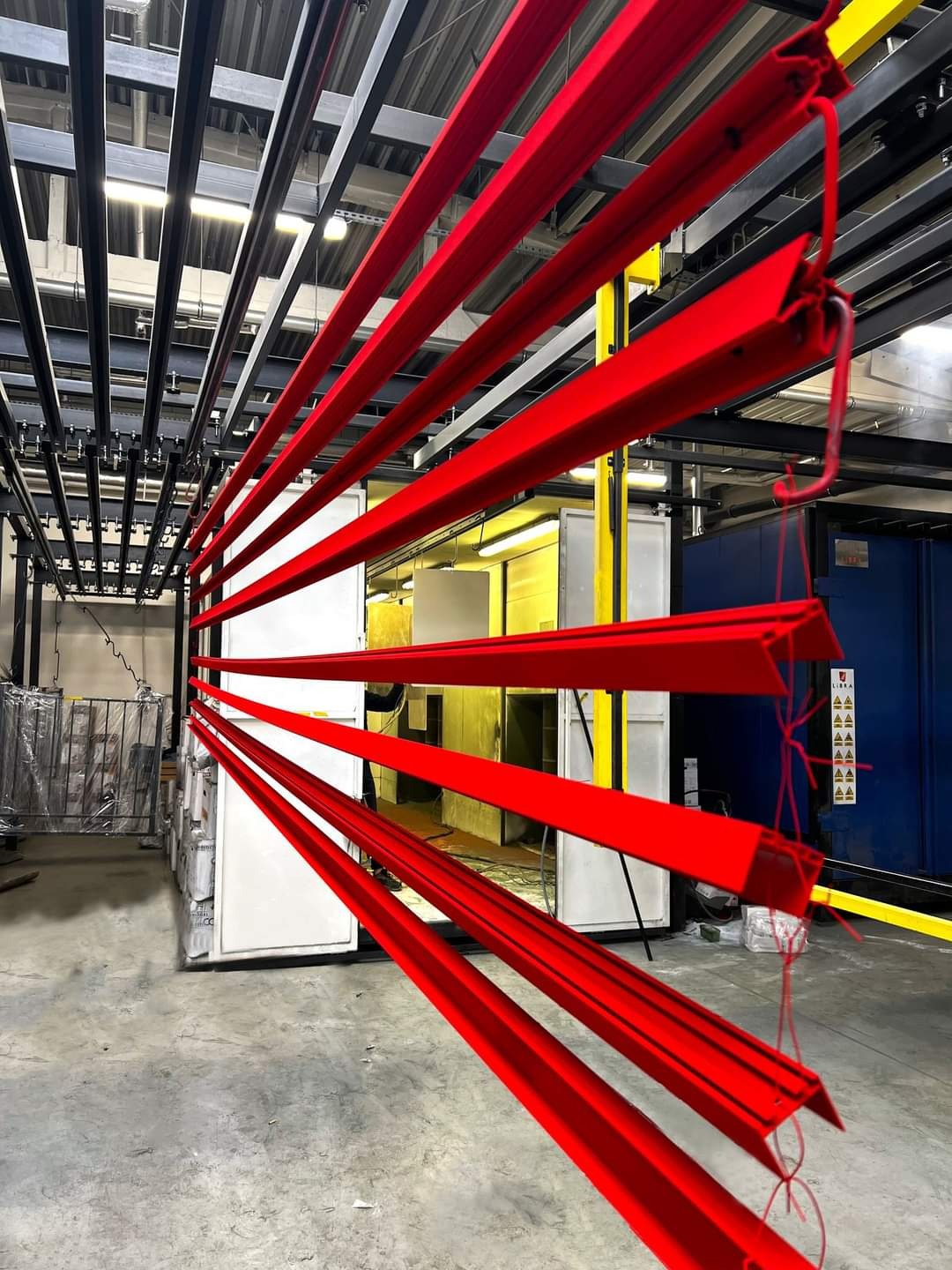 Czerwone, metalowe profile zawieszone na hakach przed piecem do malowania; widoczne elementy konstrukcji hali i podłoga przemysłowa.