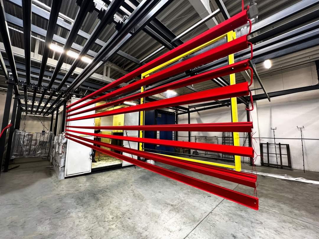Czerwone, pomalowane elementy metalowe zawieszone na prowadnicach w hali produkcyjnej, widoczny fragment pieca do utwardzania farby.