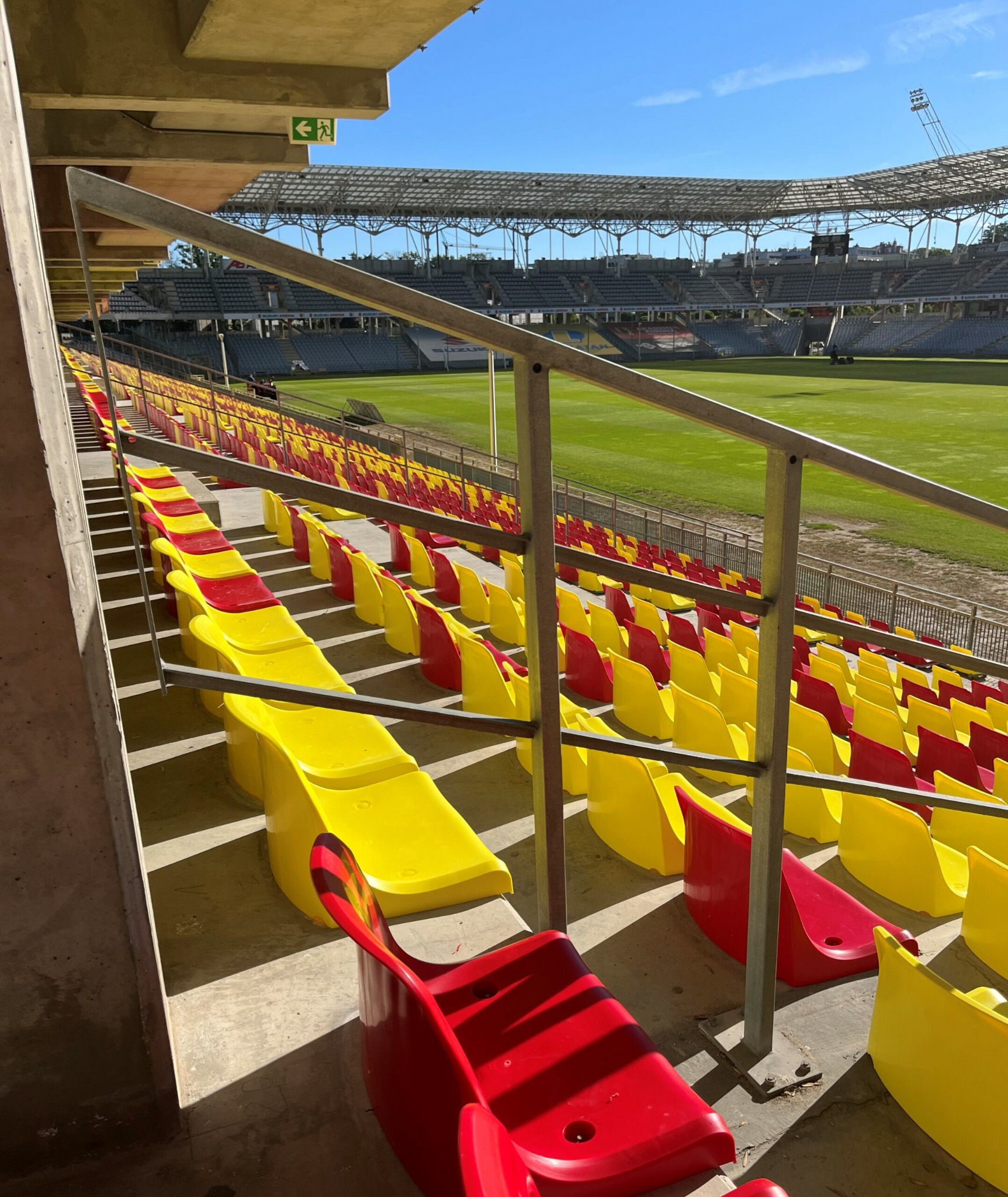 Stadionowe trybuny z rzędami plastikowych siedzeń w kolorach żółtym i czerwonym, widok z boku z perspektywy schodów, metalowa barierka w kadrze, w tle zielona murawa i zadaszenie stadionu.