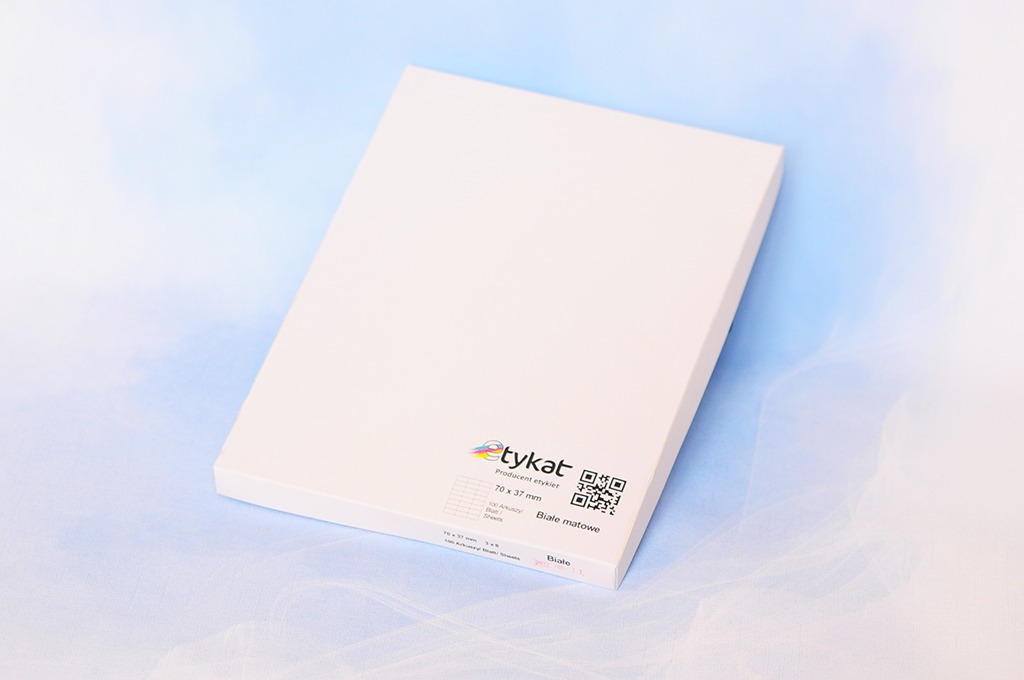 Pudełko białych, matowych naklejek samoprzylepnych formatu 70x37 mm z logo producenta 'tykat' i kodem QR, ułożone na delikatnym, błękitnym tle.