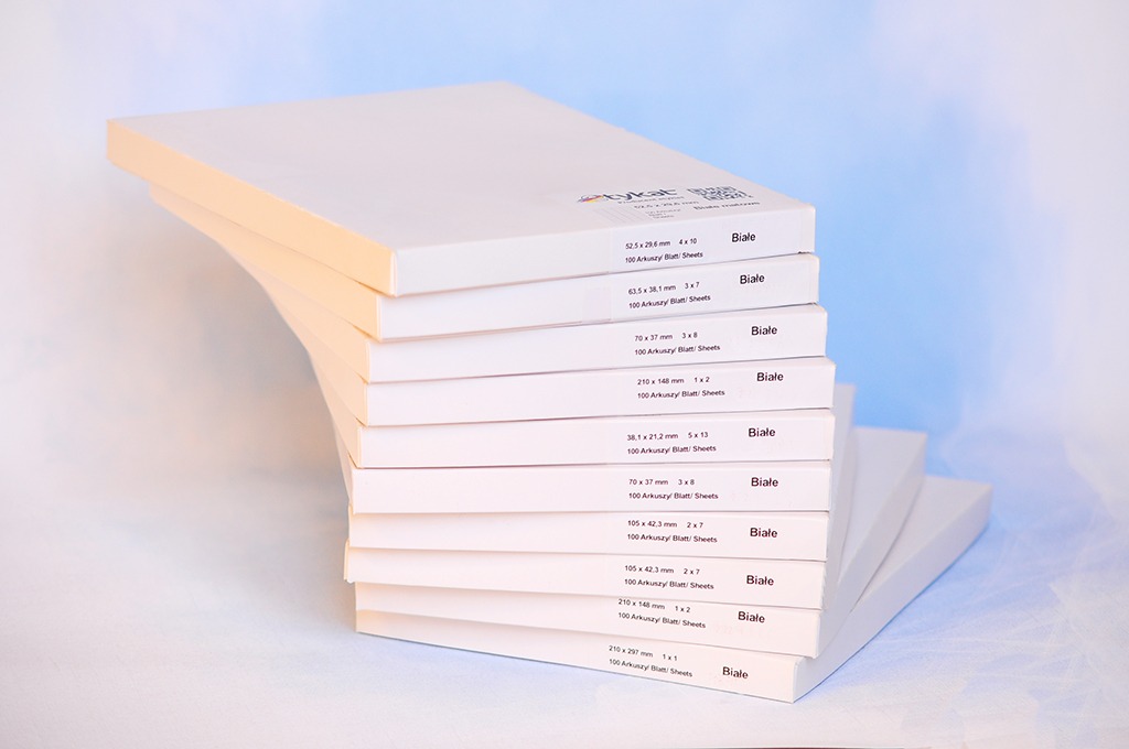 Stos białych, opakowanych arkuszy papieru z nadrukowanymi wymiarami i liczbą arkuszy, ułożonych kaskadowo na jasnym tle.
