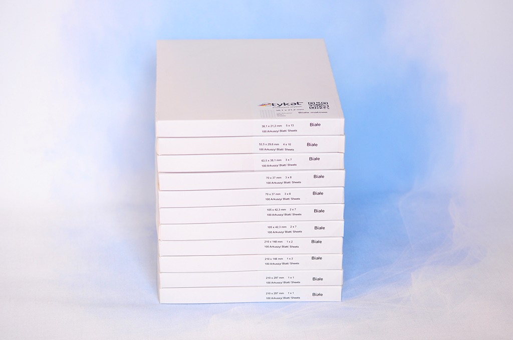 Stos białych arkuszy papieru różnych formatów, opakowanych w kartonowe pudełka z nadrukiem specyfikacji, ułożonych jeden na drugim na jasnym tle.