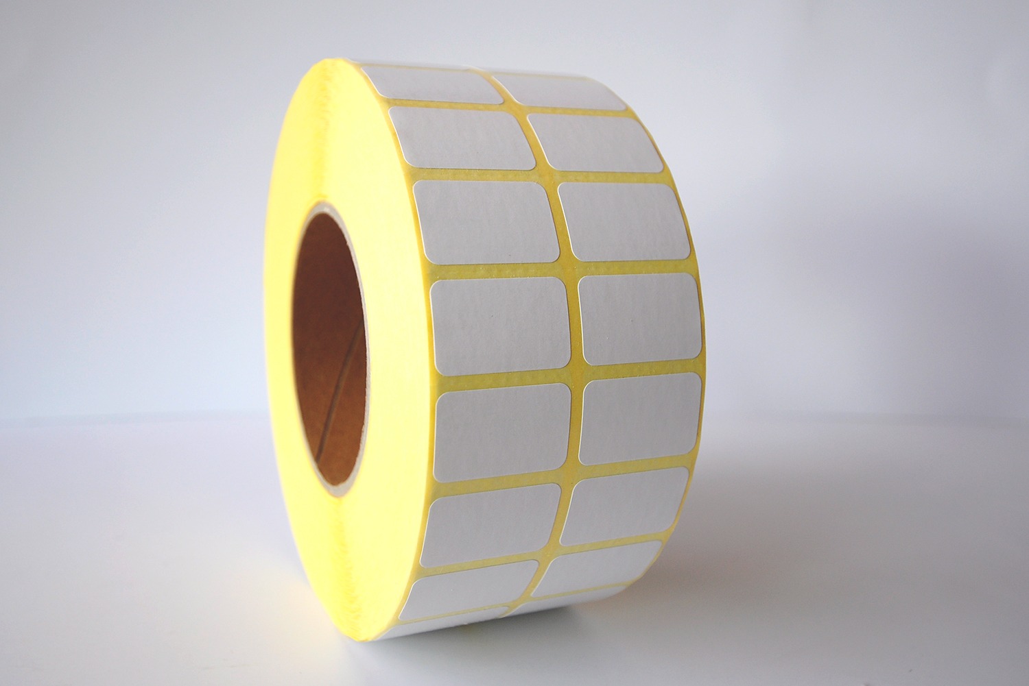 Rolka białych, prostokątnych naklejek z zaokrąglonymi rogami na żółtym podkładzie, zwinięta w rulon, widoczna pusta przestrzeń w środku rolki.