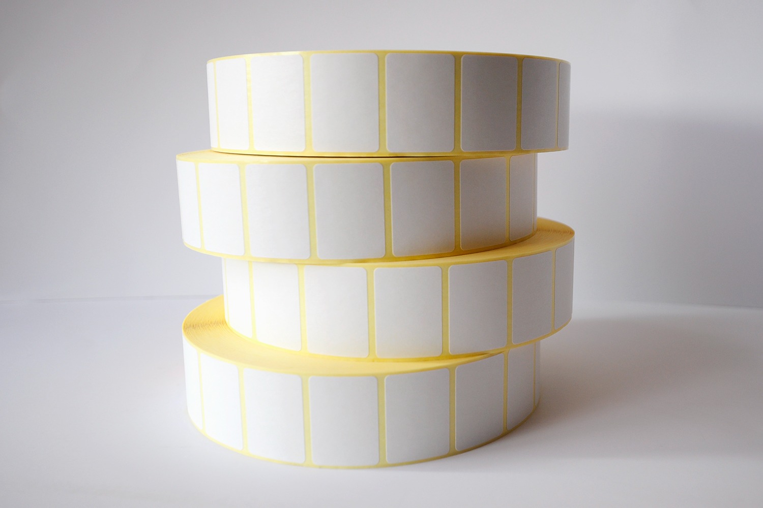 Stos czterech rolek białych, prostokątnych etykiet na żółtym podkładzie, ułożonych jedna na drugiej na jasnym tle.