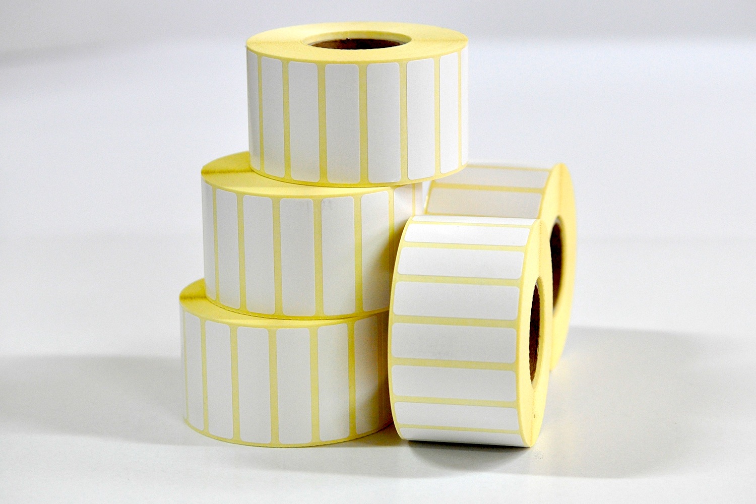Stos trzech rolek białych etykiet samoprzylepnych z żółtym podkładem, obok leży czwarta rolka, widoczne puste środki rolek.