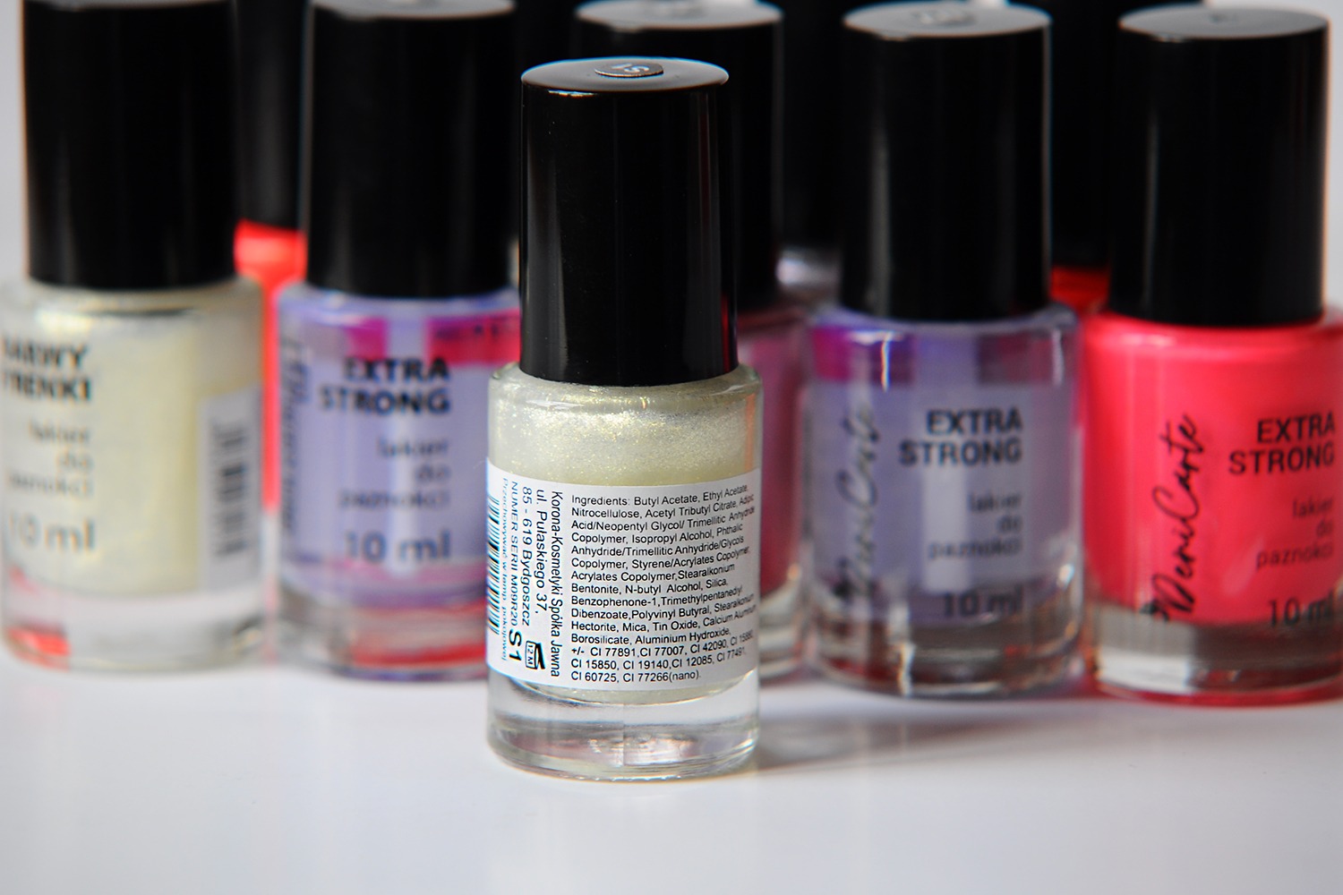 Zbliżenie na grupę lakierów do paznokci różnych kolorów i marek, z etykietami widocznymi na buteleczkach, ustawionych na białym tle.
