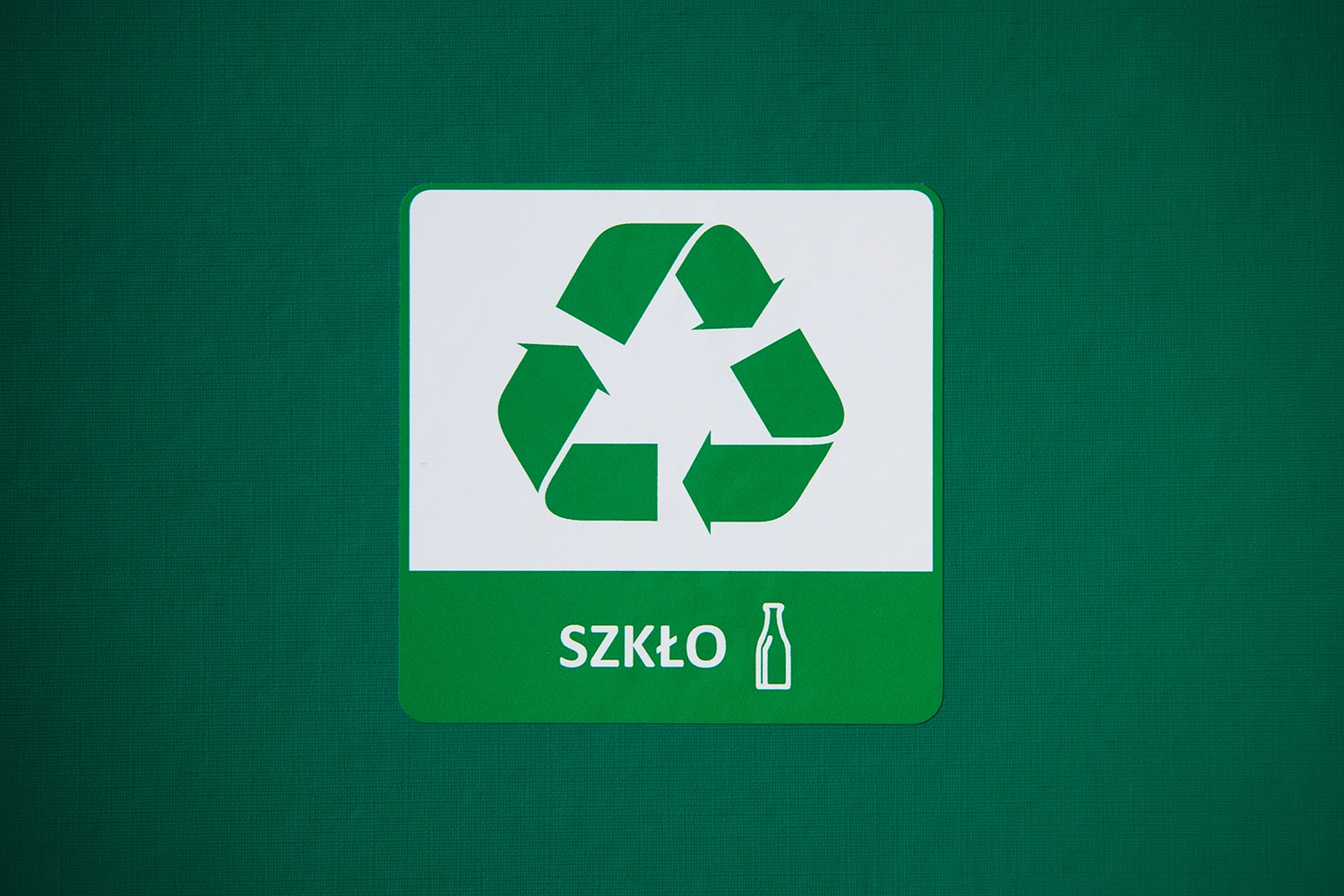 Zielony znak recyklingu szkła z symbolem butelki na białym tle, umieszczony na zielonej powierzchni.
