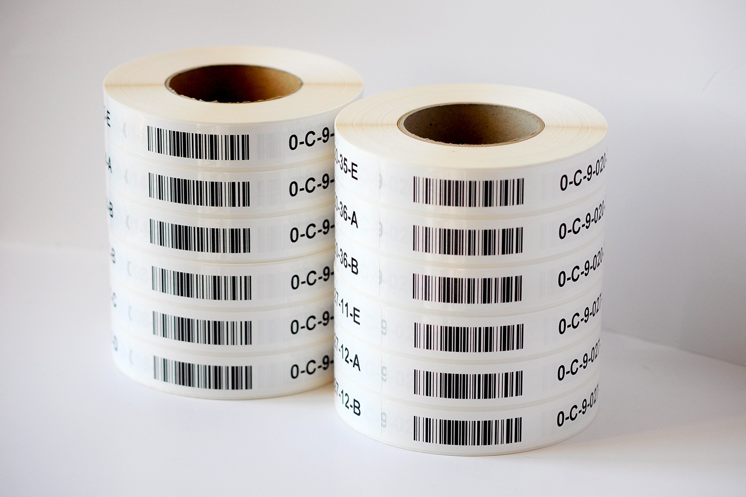 Dwa rolki białych etykiet z kodami kreskowymi i numeracją seryjną, ułożone obok siebie na jasnym tle, z widocznymi warstwami etykiet na rolkach.
