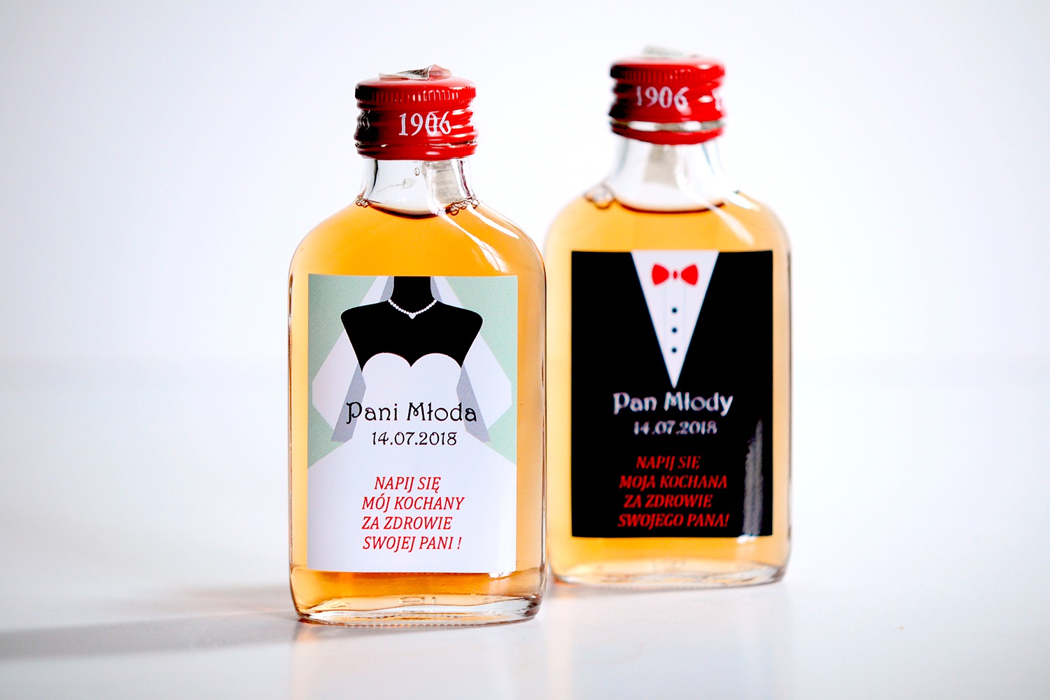 Dwie małe butelki alkoholu z niestandardowymi etykietami ślubnymi: 'Pani Młoda' z sylwetką sukni ślubnej i 'Pan Młody' z sylwetką smokingu, obie z datą 14.07.2018 i okolicznościowym tekstem...