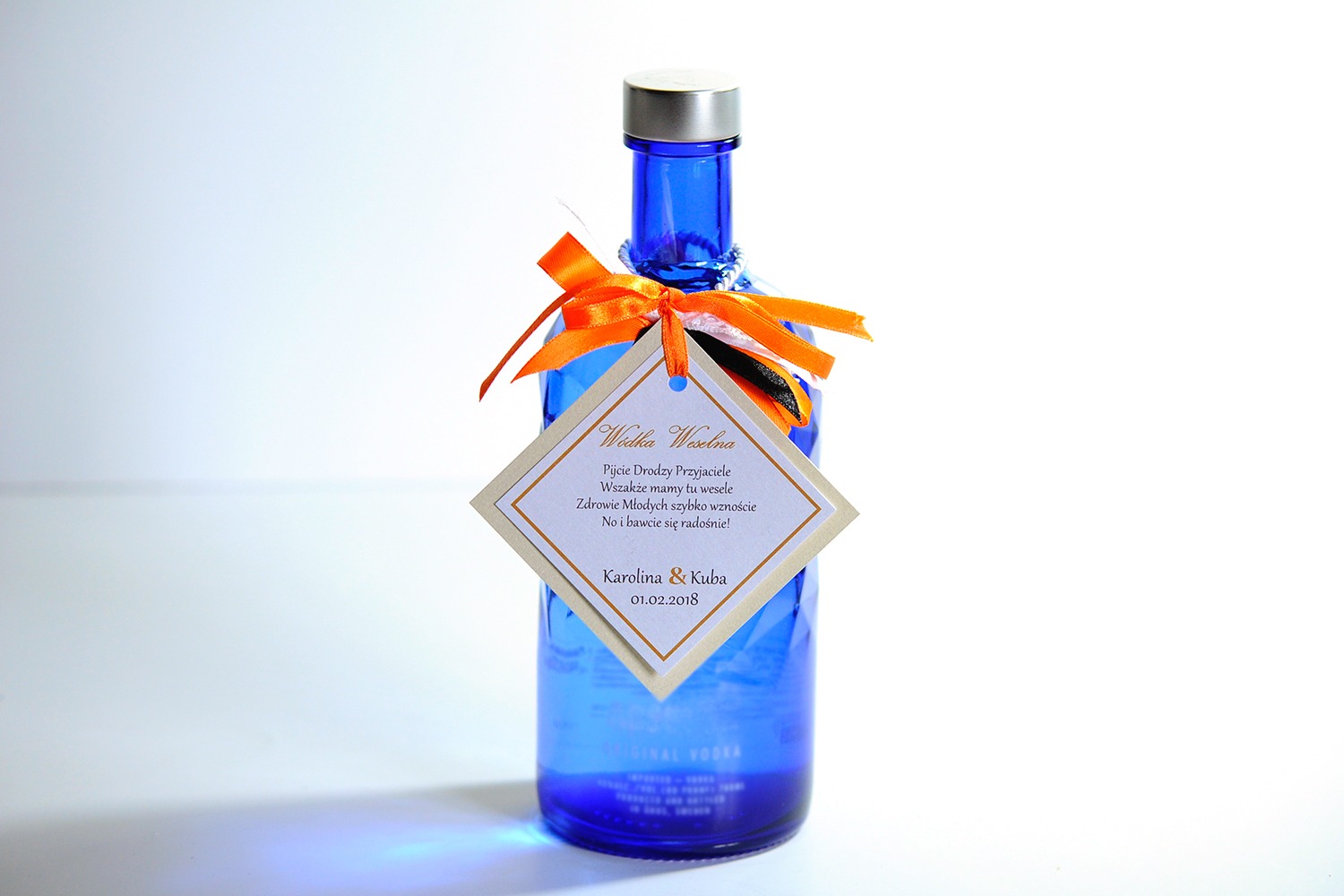 Błękitna butelka wódki weselnej z pomarańczową wstążką i spersonalizowaną etykietą z tekstem i datą ślubu Karoliny i Kuby.