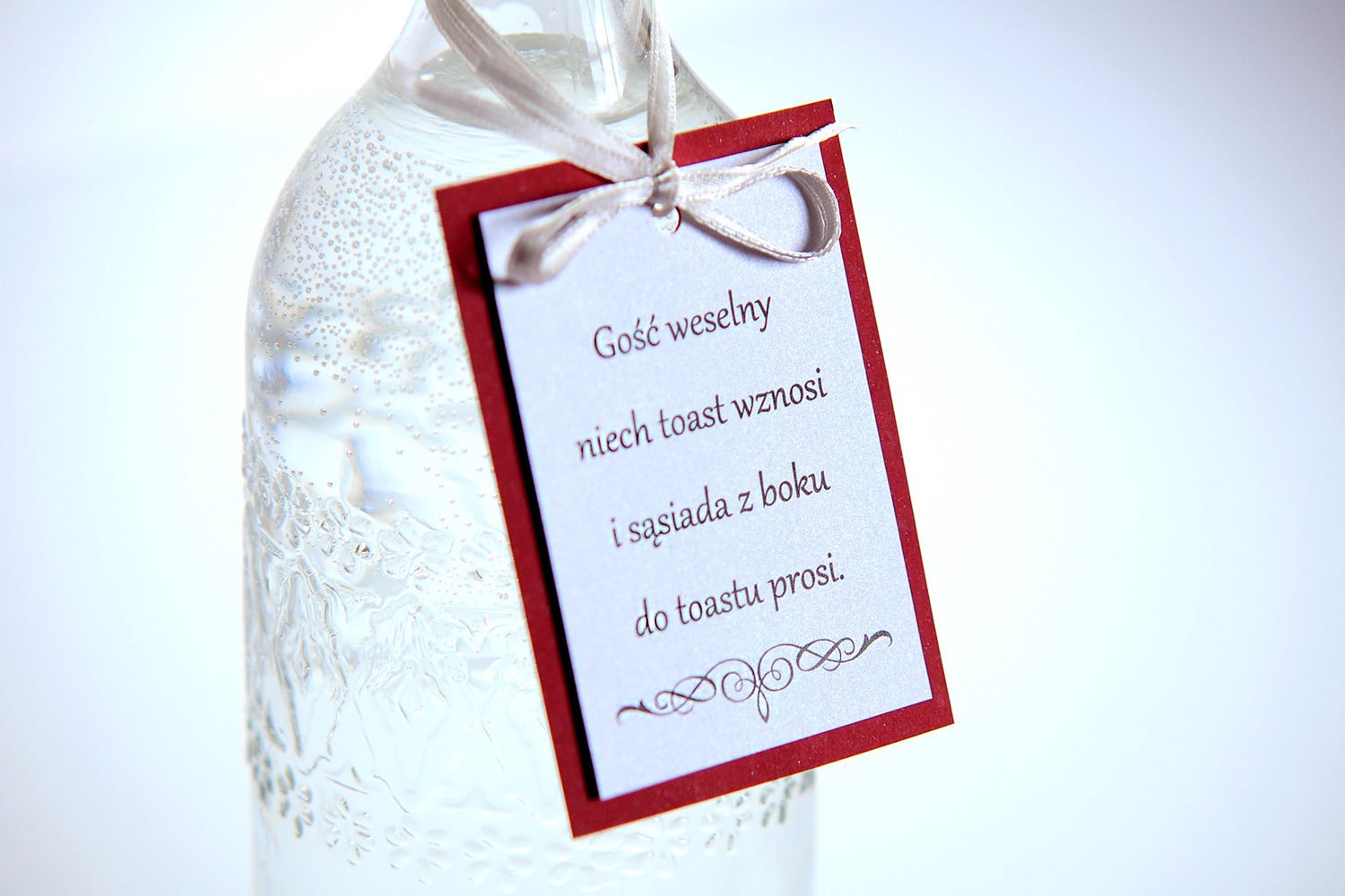 Szklana butelka z wodą gazowaną i zawieszką z tekstem 'Gość weselny niech toast wznosi i sąsiada z boku do toastu prosi' oraz ozdobnym ornamentem na dole, przewiązana satynową wstążką.