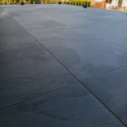 Nowoczesny taras z szarych płyt, z widokiem na dom i zieleń. Minimalistyczny design i precyzyjne ułożenie płyt tworzą elegancką przestrzeń.