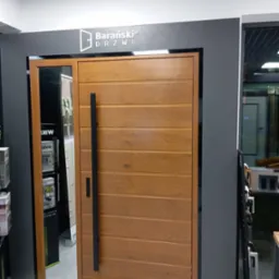 Drewniane drzwi zewnętrzne marki Barański z długim, czarnym uchwytem, wbudowane w ramę z lustrem po lewej stronie, prezentowane w salonie.