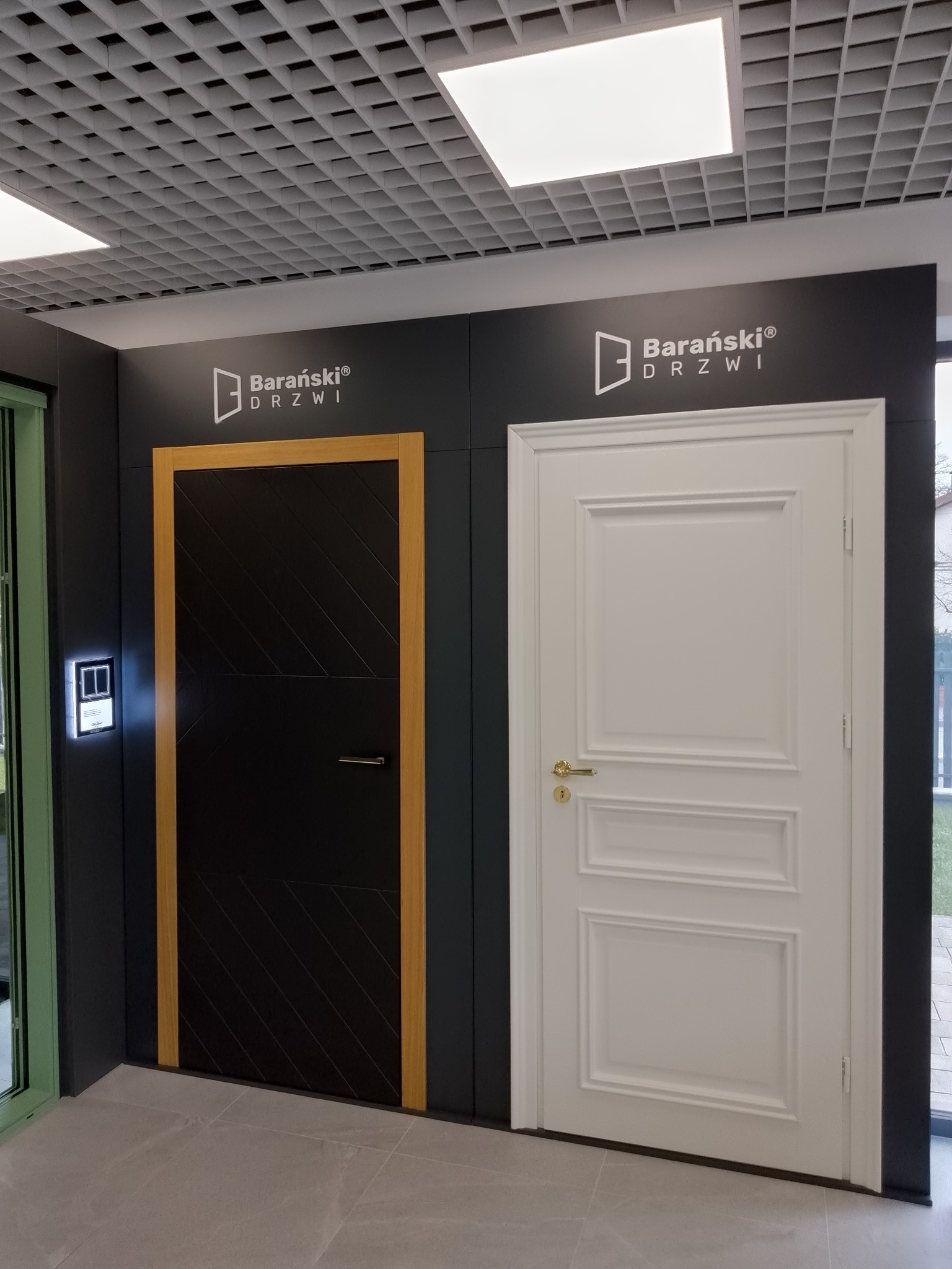Ekspozycja dwóch modeli drzwi marki Barański: czarne drzwi z drewnianą ramą i ukośnym wzorem oraz białe drzwi z ozdobnymi panelami i złotą klamką, prezentowane w salonie z podwieszanym sufitem...