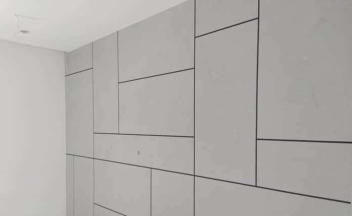Ściana wykończona panelami w odcieniach szarości o różnych kształtach, oddzielonych cienkimi, ciemnymi liniami, tworząc geometryczny wzór. Widoczny fragment sufitu i narożnik pomieszczenia.