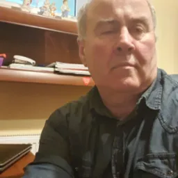 Portret mężczyzny w dżinsowej koszuli na tle p&oacute;łki z bibelotami i dokumentami.