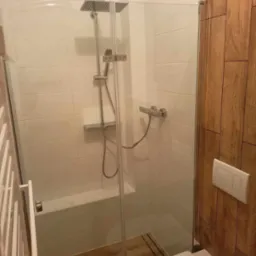 Nowoczesna łazienka z kabiną prysznicową typu walk-in, wykończona białymi płytkami i drewnianą okładziną ścienną, widoczny sedes podwieszany z białym przyciskiem spłukującym.