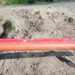 Pomarańczowa rura kanalizacyjna ustawiona na drewnianych kozłach, poziomowana czerwoną poziomicą w wykopie obok fundamentu budynku.