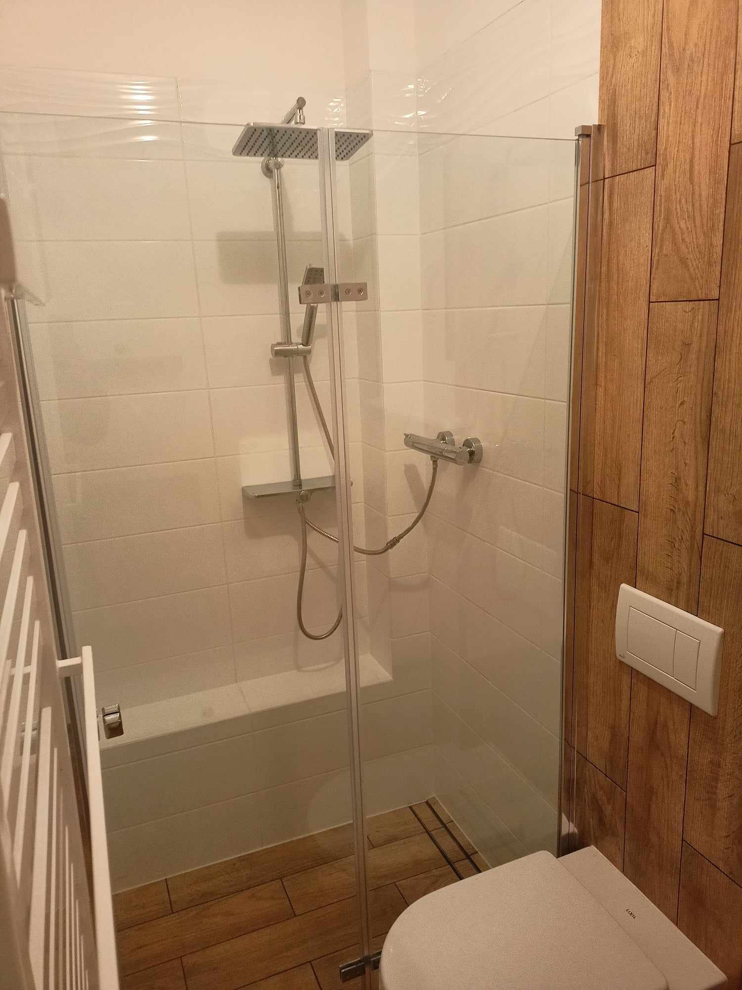 Nowoczesna łazienka z kabiną prysznicową typu walk-in, wykończona białymi płytkami i drewnianą okładziną ścienną, widoczny sedes podwieszany z białym przyciskiem spłukującym.