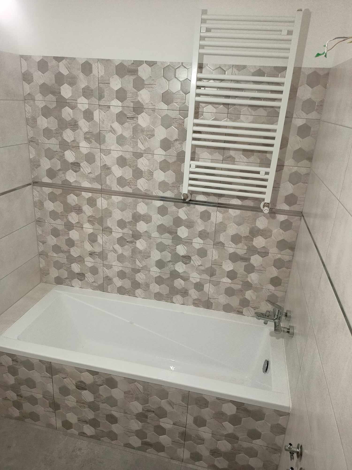 Wykończona łazienka z wanną obudowaną heksagonalnymi płytkami imitującymi drewno, biały grzejnik drabinkowy na ścianie z szarymi płytkami, chromowana bateria wannowa