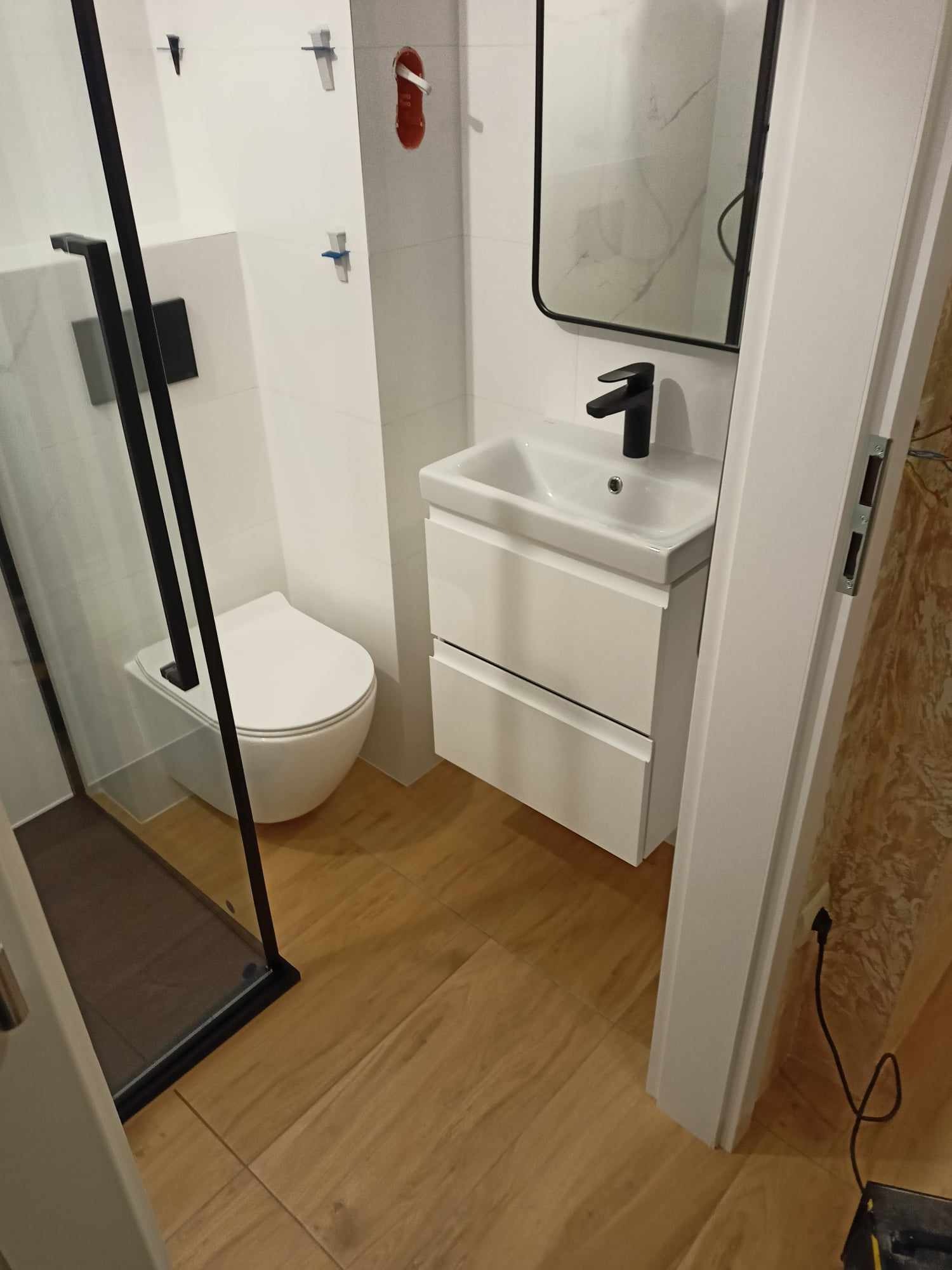 Nowoczesna łazienka z kabiną prysznicową typu walk-in z czarnymi profilami, podwieszaną toaletą, białą umywalką nablatową z czarną armaturą i lustrem w czarnej ramie, na podłodze panele drewnopodobne.