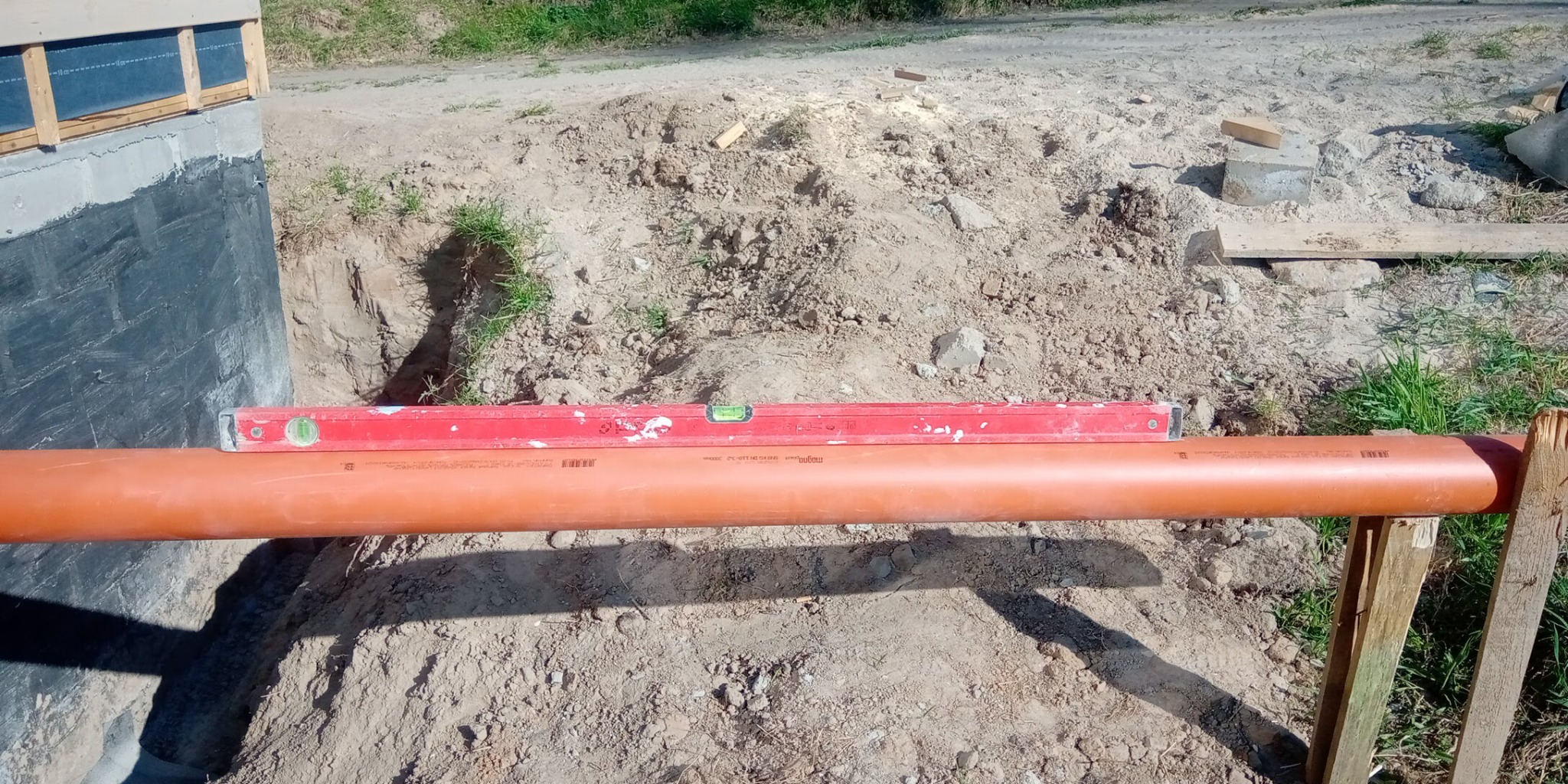 Pomarańczowa rura kanalizacyjna ustawiona na drewnianych kozłach, poziomowana czerwoną poziomicą w wykopie obok fundamentu budynku.