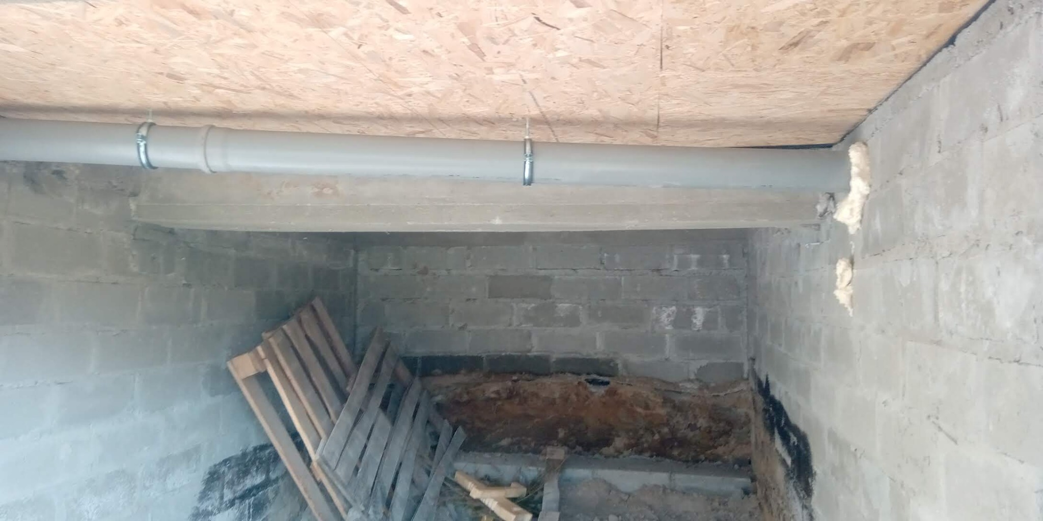Szara rura kanalizacyjna zamontowana pod stropem z płyty OSB w surowym pomieszczeniu z bloczków betonowych, widoczna izolacja pianką montażową i paleta drewniana.