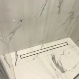 Białe marmurowe płytki w kabinie prysznicowej z odpływem liniowym, widoczne delikatne żyłki marmuru, minimalistyczny design.