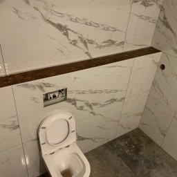 Foreman - Wykończona łazienka w skosach z białymi płytkami imitującymi marmur, brązowym pasem dekoracyjnym i częściowo widoczną toaletą. Widoczne niedokończone elementy instalacji.