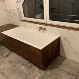 Foreman - Biała wanna obudowana brązową mozaiką w łazience z marmurowymi płytkami i szarą podłogą. Widoczna bateria w kolorze miedzi i okno.