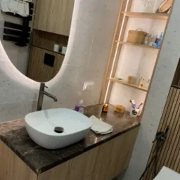 Nowoczesna łazienka z okrągłym lustrem, umywalką nablatową na blacie z ciemnego marmuru i drewnianymi półkami z oświetleniem LED.