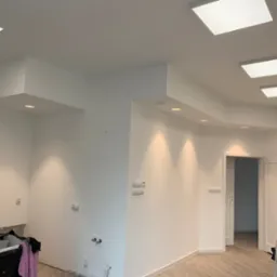 Białe wnętrze po malowaniu ścian, z widocznymi punktami świetlnymi w suficie, częściowo zabezpieczone podłoże, otwarte białe drzwi w tle i marmurowe płytki podłogowe.