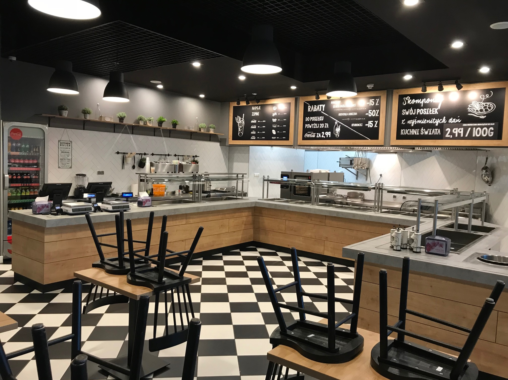 Nowoczesna aranżacja wnętrza lokalu gastronomicznego z czarno-białą podłogą i drewnianymi elementami zabudowy. Widoczne tablice z menu i oświetlenie punktowe.