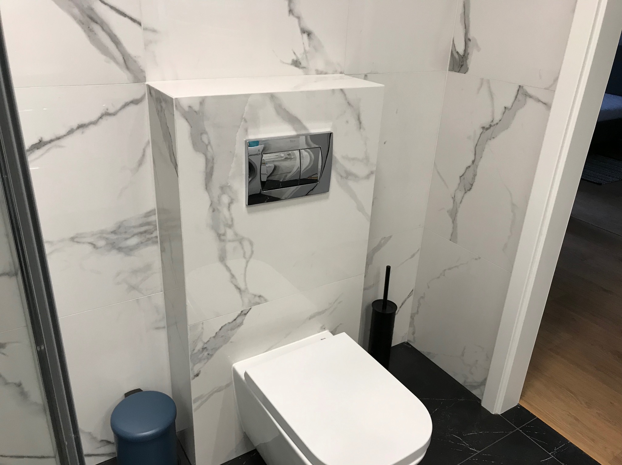 Elegancka toaleta w wykończonej łazience z białymi płytkami imitującymi marmur i czarną podłogą. Widoczny podtynkowy system spłukiwania i akcesoria.