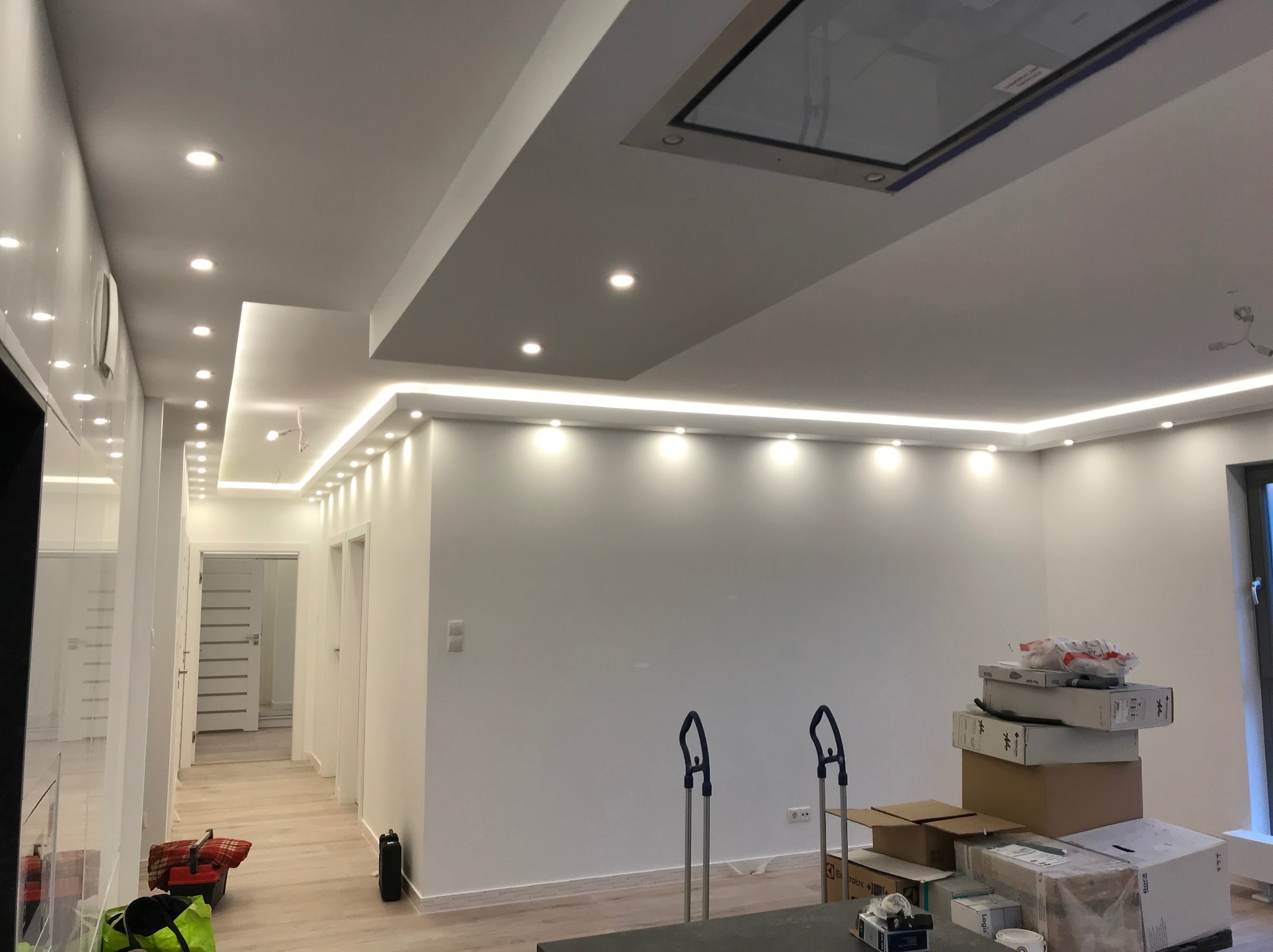 Nowoczesne wykończenie wnętrza: białe ściany, sufit z oświetleniem LED, widoczne kartony i narzędzia, jasna podłoga. Minimalistyczny design w trakcie prac.