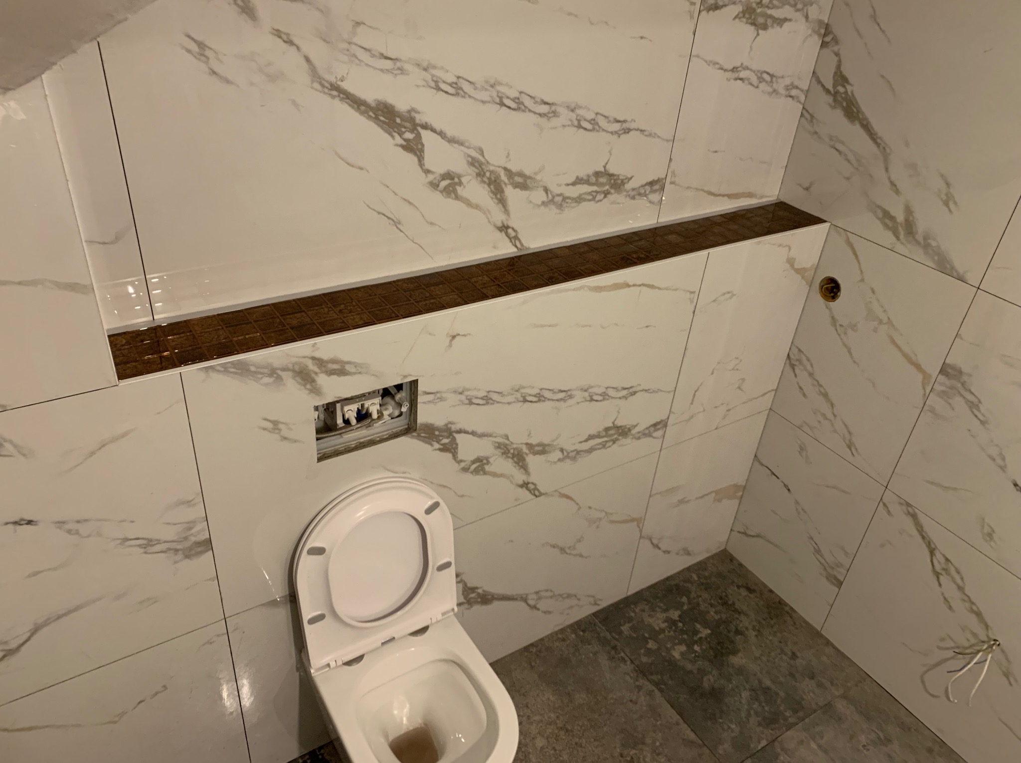 Wykończona łazienka w skosach z białymi płytkami imitującymi marmur, brązowym pasem dekoracyjnym i częściowo widoczną toaletą. Widoczne niedokończone elementy instalacji.